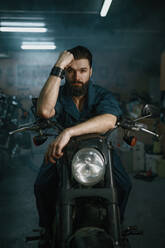 Portrait of brutal unshaven man mechanic sitting on repaired motorcycle over workshop background. Portrait of brutal man mechanic sitting on repaired motorcycle - INGF12225