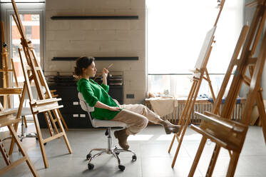 Female artist with paintbrush in hand looking seriously on her own picture while sitting on chair in art studio. Female artist with paintbrush in hand looking seriously on her own picture - INGF12228