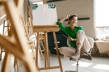 Portrait of overjoyed smiling young woman artist holding paintbrush on her nose posing for camera nearby canvas. Portrait of overjoyed smiling woman artist holding paintbrush on nose - INGF12229