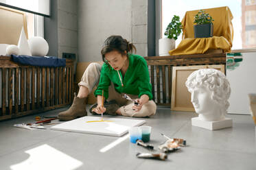 Pretty young female artist drawing picture on canvas with oil paints sitting on floor in her own art studio. Creativity in work and contemporary creative painter concept. Young female artist drawing picture sitting on floor in her own art studio - INGF12230