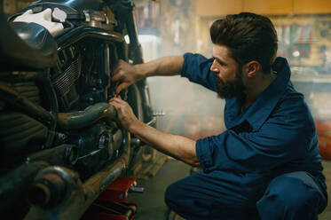 Young mechanic wearing coveralls using professional tools working on motorcycle in garage repair service. Young mechanic working on motorcycle in garage repair service - INGF12235
