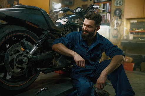 Satisfied smiling professional mechanic feeling glad looking at repaired motorcycle after good work in repair workshop. Satisfied smiling mechanic feeling glad looking at repaired motorcycle - INGF12236
