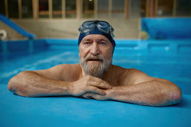 Portrait of mature senior man wearing swimming hat and goggles at pool. Grey-haired bearded male swimmer looking at camera. Concept of sport, active lifestyle and retirement. Portrait of mature senior man wearing swimming hat and goggles at pool - INGF12241