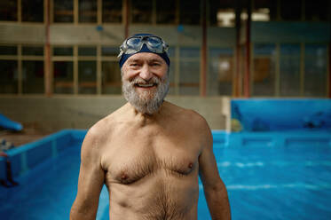 Portrait of smiling senior man with wet body in swimming pool at gym. Happy smiling athlete elderly male swimmer feeling satisfied after training looking at camera. Portrait of smiling senior man with wet body in swimming pool looking at camera - INGF12243