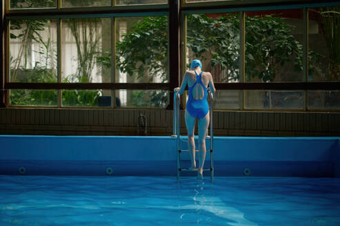 Back view on woman in wet swimsuit getting out from pool using ladder. Sport swim training finish. Back view on woman in swimsuit getting out from pool using ladder - INGF12244