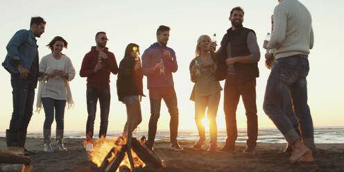 Happy Carefree Young Friends Having Fun And Drinking Beer By Bonefire On The Beach As The Sun Begins To Set - INGF12266