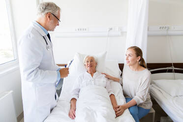 Medicine, healthcare and people concept - senior woman patient with daughter and doctor with clipboard at hospital ward. senior woman and doctor with clipboard at hospital - INGF12271