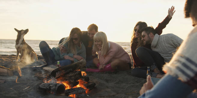 Group of friends with dog relaxing around bonfire on the beach at sunset - INGF12273