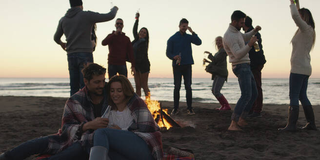 Happy Carefree Young Friends Having Fun And Drinking Beer By Bonefire On The Beach As The Sun Begins To Set - INGF12280