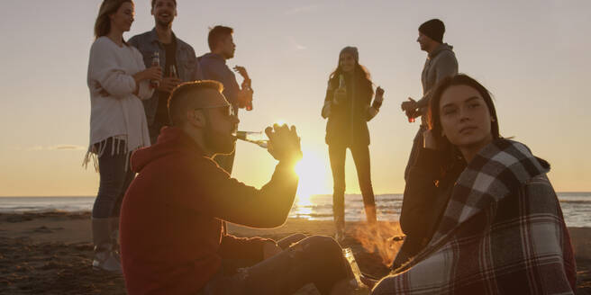 Happy Carefree Young Friends Having Fun And Drinking Beer By Bonefire On The Beach As The Sun Begins To Set - INGF12282