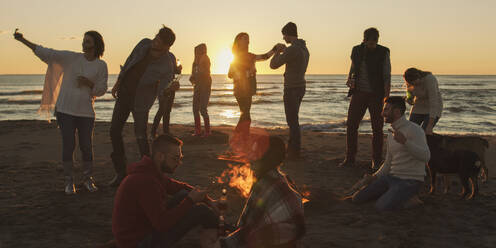 Happy Carefree Young Friends Having Fun And Drinking Beer By Bonefire On The Beach As The Sun Begins To Set - INGF12283