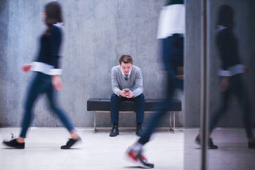 Young businessman using smart phone while sitting on the bench at busy office lobby during a break - INGF12292