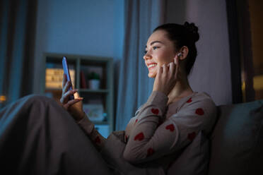 Technology, bedtime and people concept - happy smiling teenage girl with smartphone and earphones sitting in bed at home at night. teenage girl with phone and earphones in bed at night - INGF12303