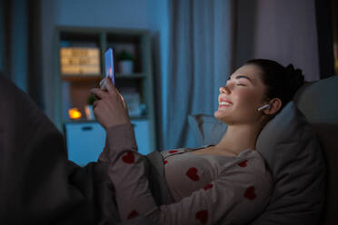 Technology, bedtime and people concept - happy teenage girl with smartphone and earphones enjoying music lying in bed at home at night. happy teenage girl with phone and earphones in bed - INGF12304