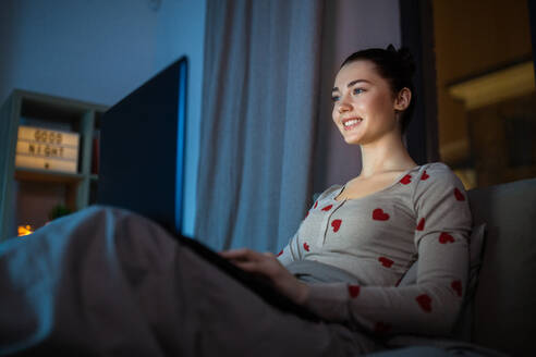 Technology, bedtime and people concept - happy smiling teenage girl with laptop computer sitting in bed at home at night. teenage girl with laptop in bed at home at night - INGF12305