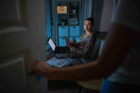 Education, technology and people concept - teenage student girl with notebook and laptop computer learning in bed at home at night and mother entering room. student girl with laptop learning at night - INGF12306