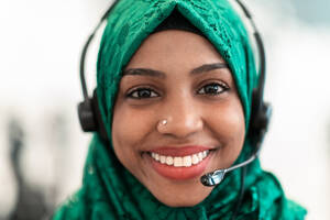 Afro Muslim female with green hijab scarf customer representative businesswoman with phone headset helping and supporting online with the customer in a modern call center. High-quality photo. Afro Muslim female with green hijab scarf customer representative business woman with phone headset helping and supporting online with customer in modern call centre - INGF12312