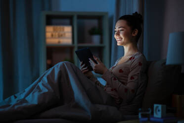 Technology, bedtime and people concept - happy smiling teenage girl tablet pc computer sitting in bed at home at night. teenage girl with tablet pc in bed at night - INGF12313