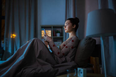 Technology, bedtime and rest concept - teenage girl in pajamas with smartphone sitting in bed at night. teenage girl in pajamas with phone in bed at night - INGF12314