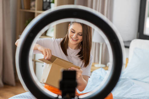 Blogging, technology and people concept - happy smiling girl blogger with ring light and smartphone unpacking parcel box at home. happy girl blogger unpacking parcel box at home - INGF12316