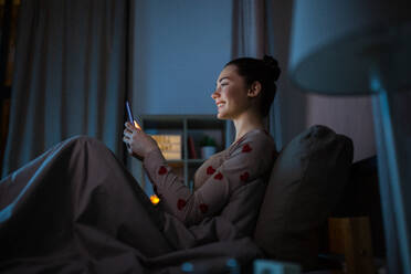 Technology, bedtime and rest concept - happy smiling teenage girl in pajamas with smartphone sitting in bed at night. teenage girl in pajamas with phone in bed at night - INGF12323