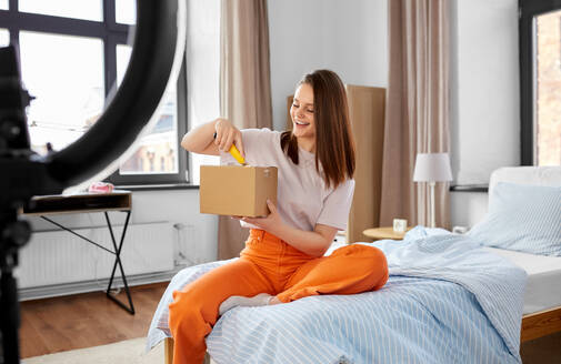 Blogging, technology and people concept - happy smiling girl blogger with ring light and smartphone unpacking parcel box with knife at home. happy girl blogger unpacking parcel box at home - INGF12329