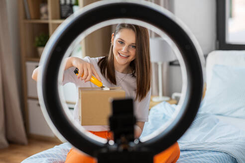 Blogging, technology and people concept - happy smiling girl blogger with ring light and smartphone unpacking parcel box with knife at home. happy girl blogger unpacking parcel box at home - INGF12330
