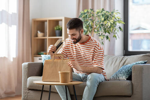 Consumption, eating and people concept - smiling man unpacking takeaway food in paper bag at home. smiling man unpacking takeaway food at home - INGF12342