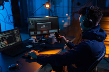 Cybercrime, hacking and technology concept - male hacker with smartphone using computer virus program for cyber attack in dark room. hacker with smartphone and computers in dark room - INGF12345