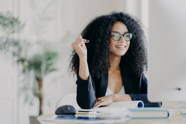 Pretty Afro American woman looks positively at display of computer, writes down information from internet, wears optical glasses and black suit, sits at desktop with necessary things for work - INGF12346