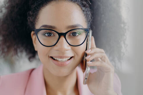 Close up portrait of happy dark skinned businesswoman holds mobile phone near ear, calls business partner, has charming smile, wears optical glasses, has curly bushy hair, discusses working issues - INGF12347