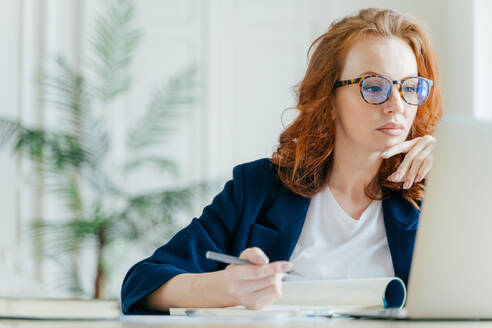 Serious experienced female coach makes records in notepad, focused at screen of laptop computer, has long red hair, wears transparent glasses, watches webinar online. Business and job concept - INGF12348