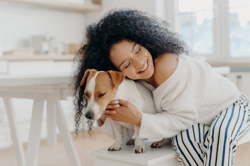 Lovely young curly African American woman embraces beloved pedigree dog with love, has gentle smile, wears stylish clothing, poses against home background in modern apartment, expresses care - INGF12349