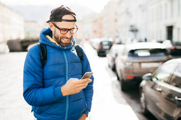 Young bearded hipster guy in blue anorak and cap holding smartphone answering call having happy expression isolated over big city background. Stylish man using cell phone while standing on street - INGF12352