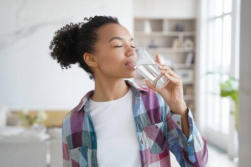 African american woman enjoy drinking pure crystal filtered potable water. Thirsty female with eyes closed swallow mineral aqua from glass at home kitchen. Thirst, healthy lifestyle, body care concept. African american young girl enjoy drinking pure filtered water from glass at home. Healthy lifestyle - INGF12363