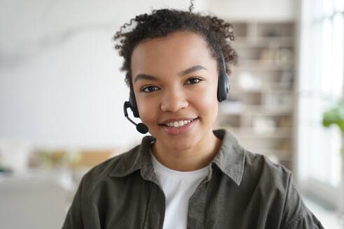 Lächelnde gemischtrassige junge Studentin mit Headset und Mikrofon, die online eine Fremdsprache lernt, freundliches afroamerikanisches Teenager-Mädchen mit Kopfhörern, das in die Kamera schaut. Fernunterricht, E-Learning. Gemischtrassige Studentin mit Headset, die online lernt und in die Kamera schaut. Fernunterricht, E-Learning - INGF12364