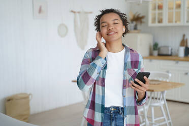 Music listening in airpods on mobile phone. Teenage girl enjoying music in wireless earphones. Afro young woman relaxing at home. Having fun and recreation alone. Music service website or app concept.. Teenage girl enjoying music in wireless earphones. Having fun and recreation concept. - INGF12365