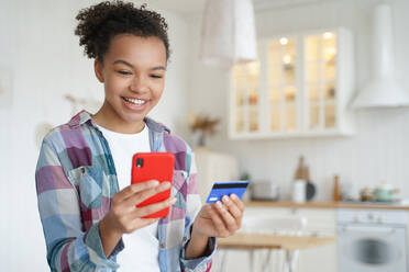 Happy hispanic young woman is purchasing online with mobile phone and credit card. Smiling teenage girl spends money from home. Online shopping, internet banking. Business and e-commerce.. Happy young woman is purchasing online with mobile phone and credit card. Internet banking. - INGF12387