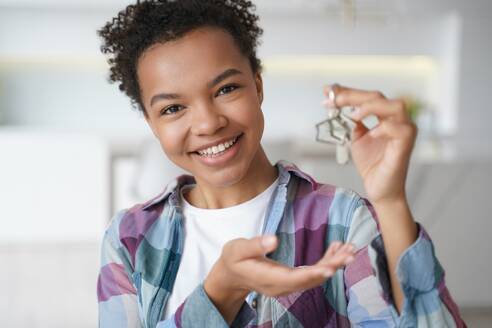 Happy young biracial girl showing key to new home, first house, apartment, looking at camera. Smiling mixed race female houseowner renter tenant holding bunch of keys. Real estate agency advertisement. Smiling young biracial girl houseowner tenant holds house key to new home. Real estate service - INGF12388