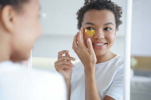 Mirror reflection of young hispanic lady. Happy afro girl is applying eye patches to her face. Skin moisturizer with collagen serum. Anti age patches. Morning skincare. Beauty routine of teenage girl.. Mirror reflection of young hispanic lady with eye patches. Skin moisturizer with collagen serum. - INGF12399