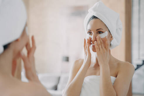 Girl applies eye patches and looking into mirror. Attractive european woman wrapped in towel after bathing. Young hispanic lady takes shower at home. Relaxation at spa resort. Beauty routine at home.. Girl applies eye patches and looking into mirror. Attractive woman wrapped in towel after bathing. - INGF12401