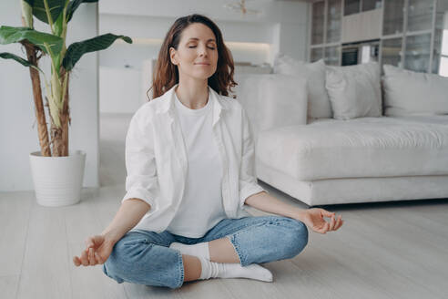 Young caucasian woman is sitting in lotus asana. Girl is practicing yoga and meditation on floor at home. Morning gymnastics. Harmony and mind relax concept. Scandinavian interior of living room.. Caucasian woman is sitting in lotus asana at home in morning. Scandinavian interior of living room. - INGF12402