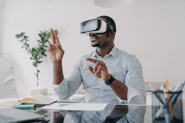 Businessman in vr headset clicks virtual buttons. Freelancer is working on design project. African american man in futuristic vr headset at home. Modern digital technology for business and creativity.. Businessman in vr headset clicks virtual buttons. Modern technology for business and creativity. - INGF12404