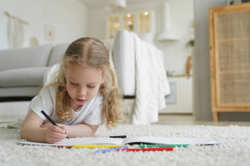 Talented little preschool child girl painting lying on warm floor in living room at home. Caucasian kid girl drawing with colored pencils playing spending leisure time alone. Children’s creativity. Talented preschool kid girl painting with colored pencils, lying on floor at home. Child creativity - INGF12406