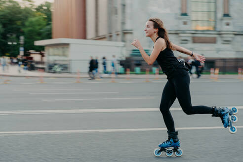 Outdoor photo of active dark haired woman in good physical shape enjoys extreme sport rides on rollers along road at busy street relaxes during weekend in open air. Healthy lifestyle concept - INGF12407