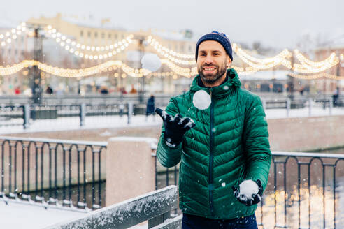 Outdoor shot of happy smiling bearded male in warm jacket and hat plays snowballs as stands against beautiful lights, being in good mood, has joyful expression. People, winter and fun concept - INGF12410