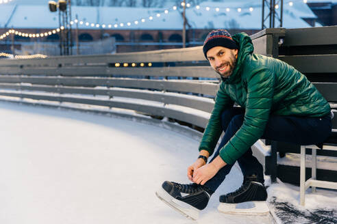 Cheerful man with appealing appearance laces up skates, sitts on ice arena, wants to go skating with friends, poses at camera. Happy bearded man enjoys having active lifestyle. Winter and season - INGF12411