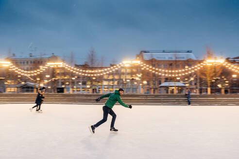 Active sorty male has fun in outdoor park on ice rink decorated with garlands, shows his talents of skating, makes fast movements on skates, being confident, enjoys winter outdoor activities - INGF12412