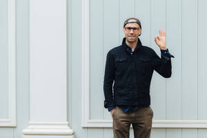 Indoor shot of handsome cheerful unshaven male shows okay sign, wears stylish shirt, cap and trousers, keeps hand in pocket, poses against wall, satisfied with something, Body language and people - INGF12415