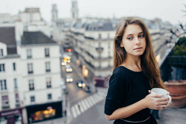 Pretty dreamy woman with mug standing on balcony and looking away in Paris, France. . Dreamy woman with mug on balcony - INGF12418